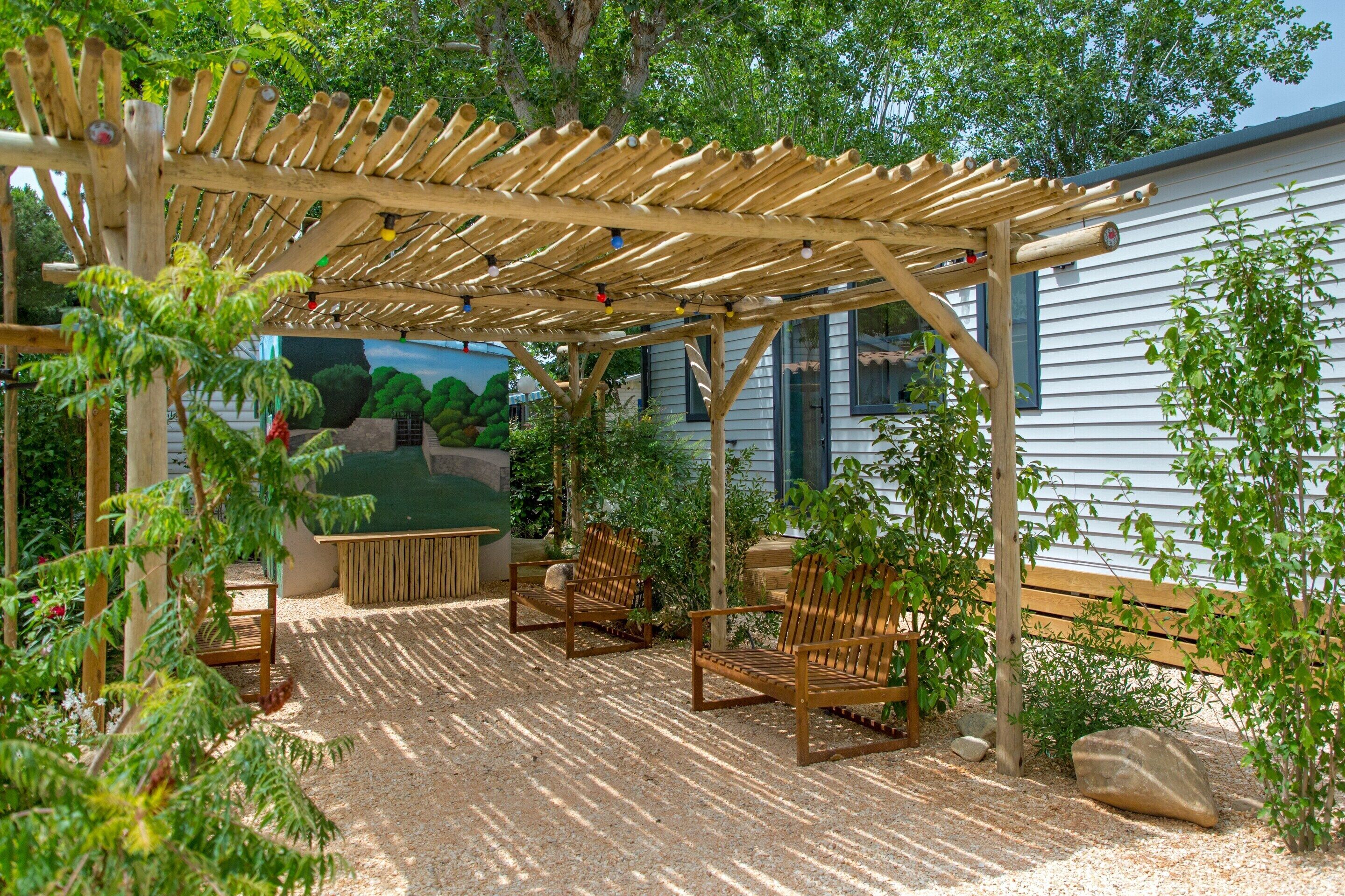 Uma pérgola de madeira com assentos, rodeada por cascalho e vegetação.