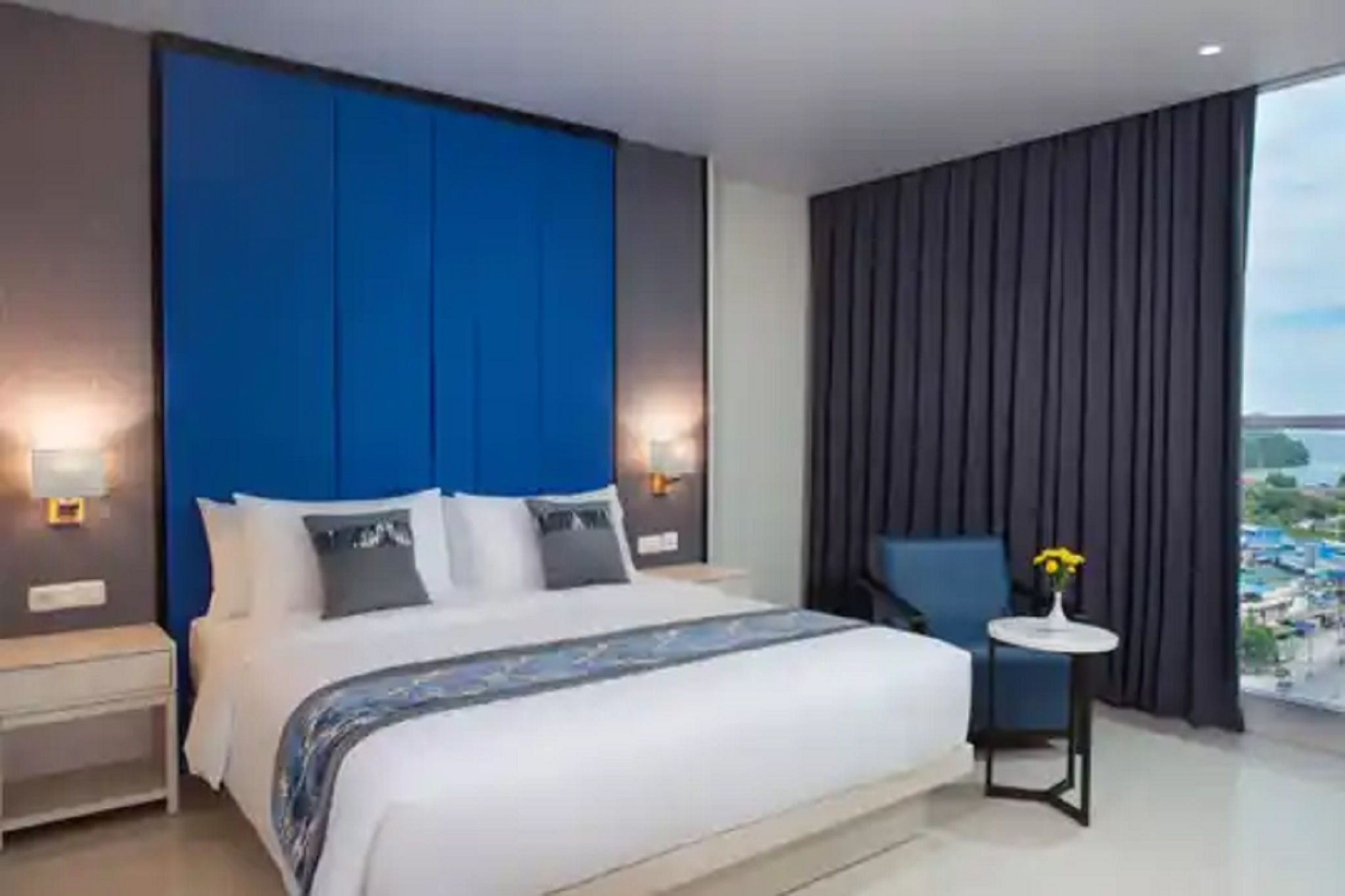 A modern hotel room with a large bed, a blue accent wall, a view of the sea, and a small table with a vase of flowers.