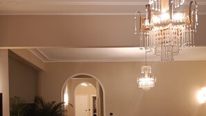Un vaste salon agrémenté d’un grand canapé en cuir marron, d’une table basse recouverte d’une nappe à motifs floraux et d’un lustre. Des plantes en pot sont disposées le long des murs, et un couloir mène à une autre pièce.