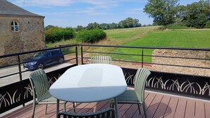 A deck with a table and chairs, overlooking a grassy field and a stone building. - Logis de la Provosterie - CROCUS (Ahuille)