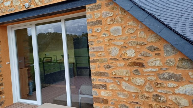 A stone building with a large glass door leading to an outdoor area with a wooden deck and a chair. - Logis de la Provosterie - CROCUS (Ahuille)