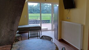A room with a table and chairs, a TV mounted on the wall, and a sliding glass door leading to an outdoor area with a patio. - Logis de la Provosterie - CROCUS (Ahuille)