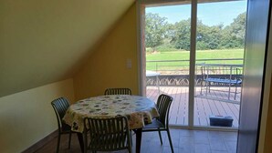 A room with a round table and four chairs, a sliding glass door leading to an outdoor deck with a metal railing. - Logis de la Provosterie - CROCUS (Ahuille)
