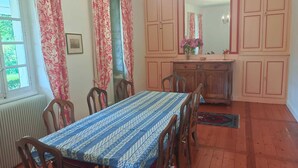Una sala menjador amb una taula preparada per a quatre, cadires de fusta, un radiador, una finestra amb cortines i un mirall sobre la porta.