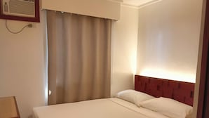 Une chambre d’hôtel avec un lit simple, une table de chevet en bois, un climatiseur fixé au mur et une fenêtre avec des rideaux.