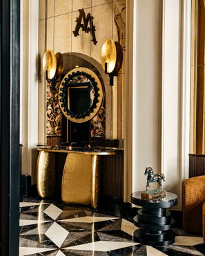 Un lobby lujoso con suelo de mármol, un mostrador de recepción con detalles dorados, un espejo decorativo y un aplique en la pared.