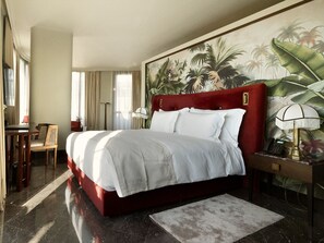 Habitación de hotel con una cama grande, un escritorio, una silla, una lámpara y un cuadro de follaje tropical en la pared.