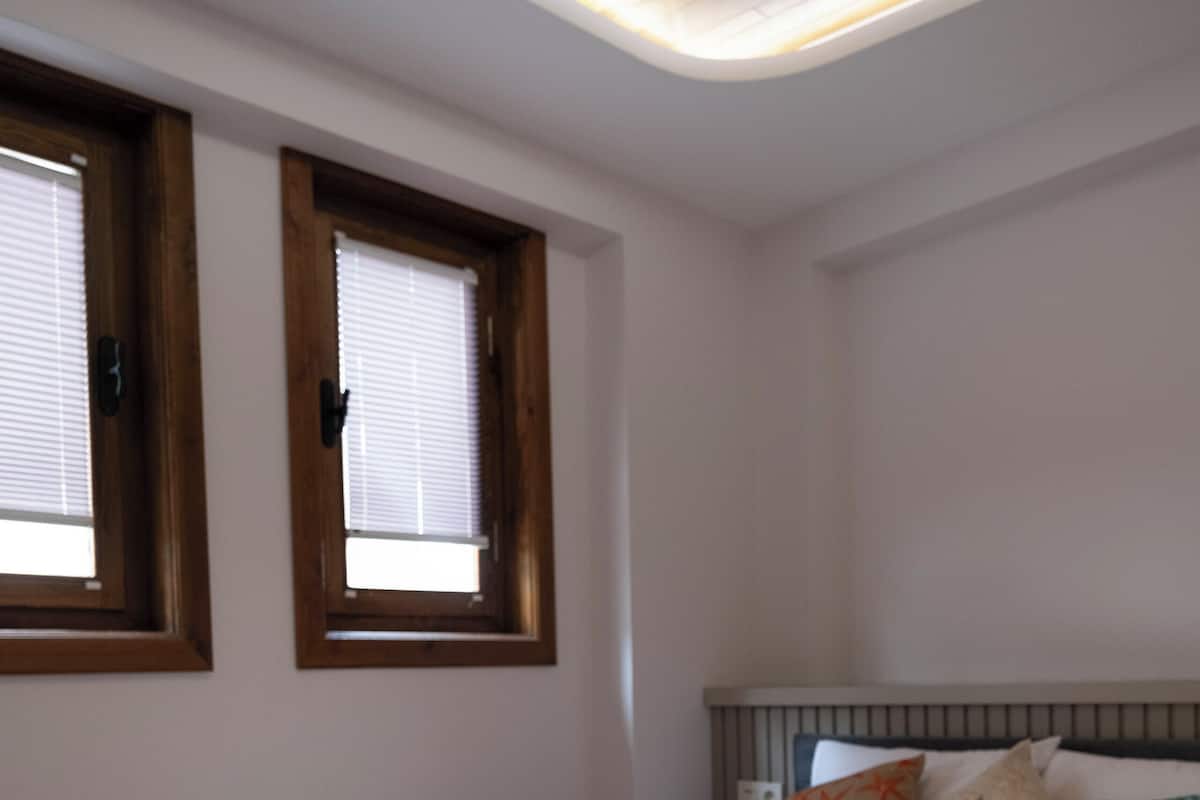 Un dormitorio con cama, un banco, dos ventanas y una lámpara de techo.