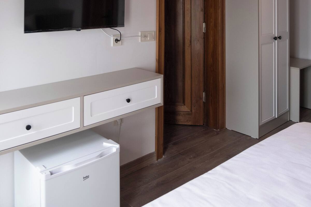 Un dormitorio con techo de madera, un televisor de pantalla plana montado en la pared, una cama blanca, un refrigerador blanco pequeño y un armario.