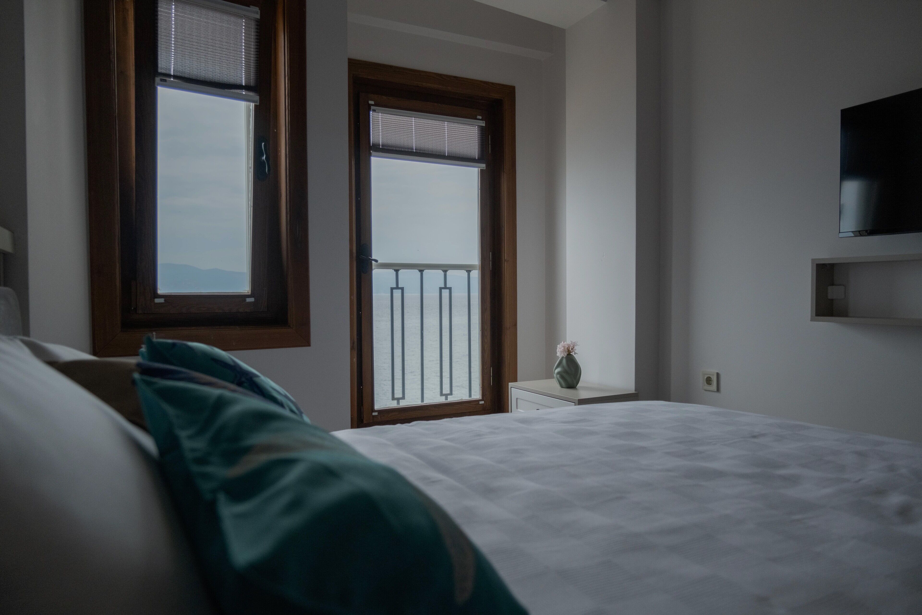 A bedroom with a bed, two windows, a small bedside table with a vase, and a mounted TV.