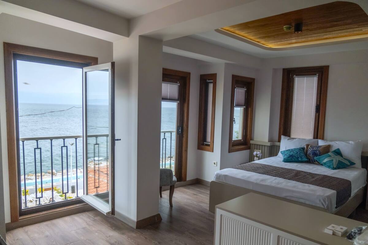 Un dormitorio con un gran ventanal con vistas a la playa, una cama con muchas almohadas y una mesita pequeña.