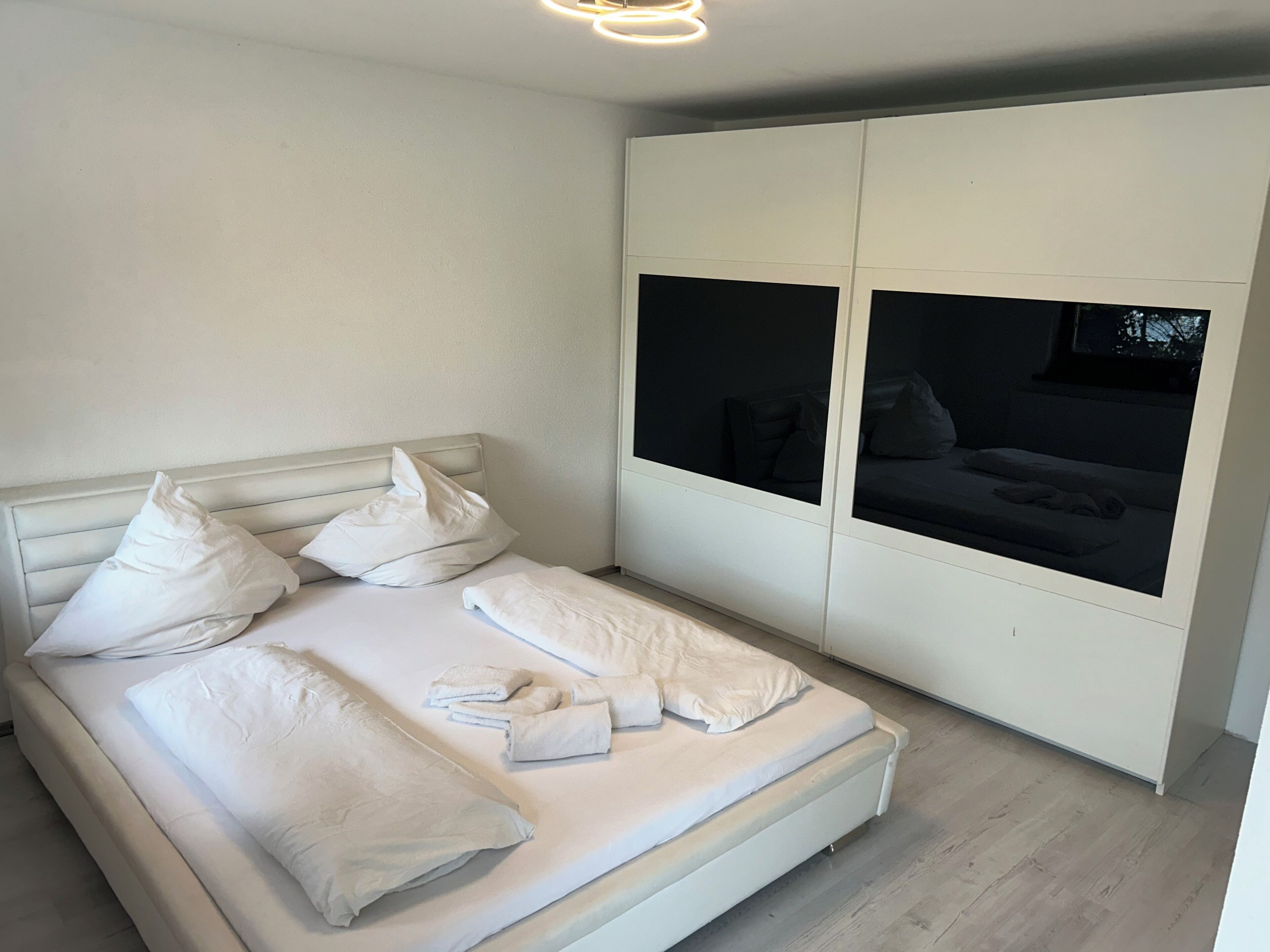 Ein ordentlich bezogenes Bett mit weißen Leinen und Kissen, ein weißer Kleiderschrank mit Spiegel und ein Blick in einen dunklen Raum durch den Spiegel.