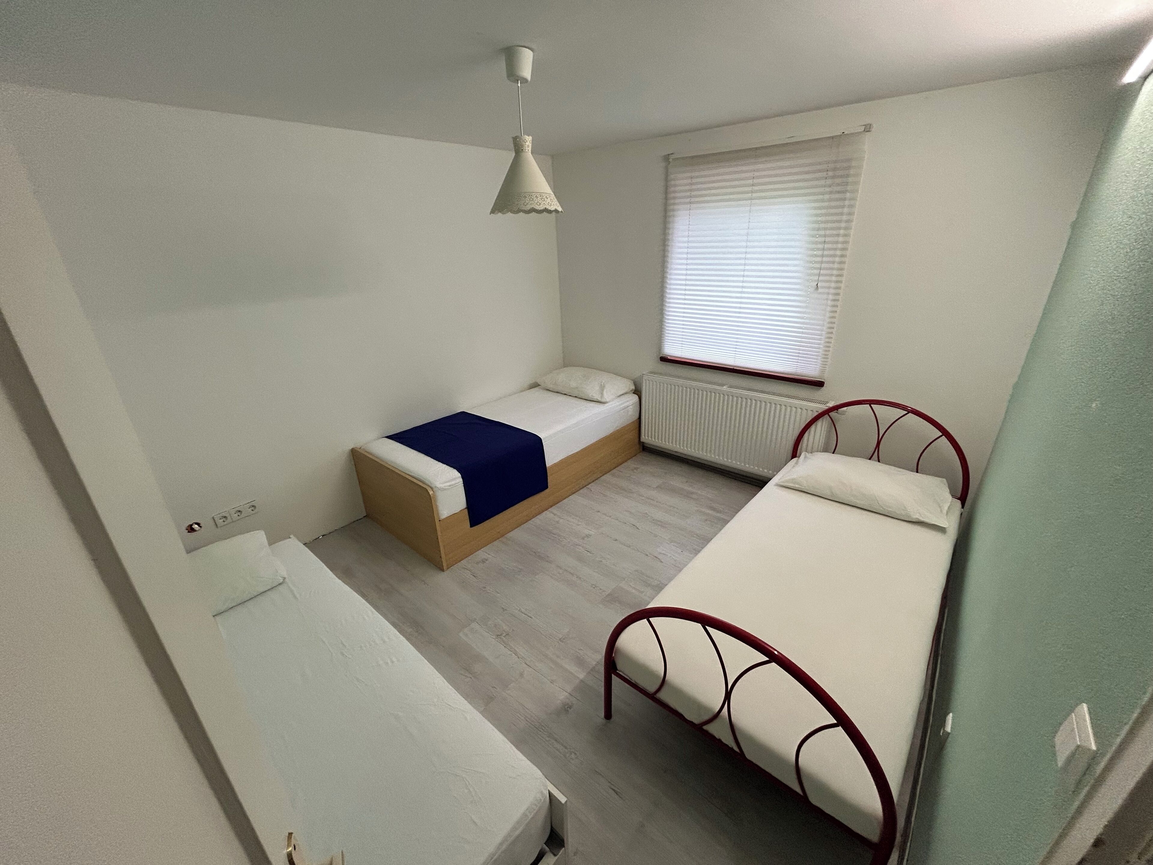 A small, well-lit room with a single bed, a bedside table, and a window with blinds.