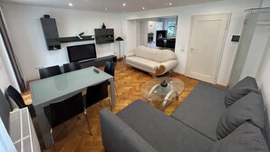 A modern living room with a sofa, a glass coffee table, a dining table with chairs, a TV, and a kitchen area in the background. - Cozy terraced house for up to 8 people1 minute from the trade fair (Offenburg)