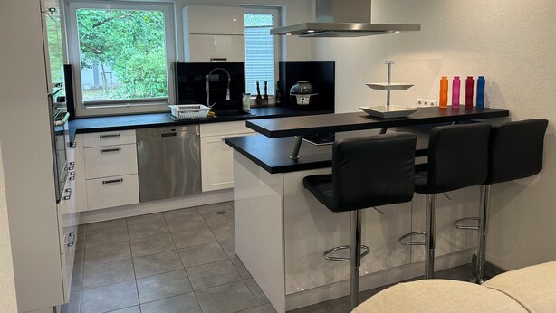 A modern kitchen with white cabinets, a stainless steel range hood, a black countertop, and a bar area with black bar stools. - Cozy terraced house for up to 8 people1 minute from the trade fair (Offenburg)