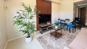 Un salon moderne avec une grande plante en pot, un téléviseur à écran plat, une table à manger avec des chaises et un luminaire suspendu au plafond.