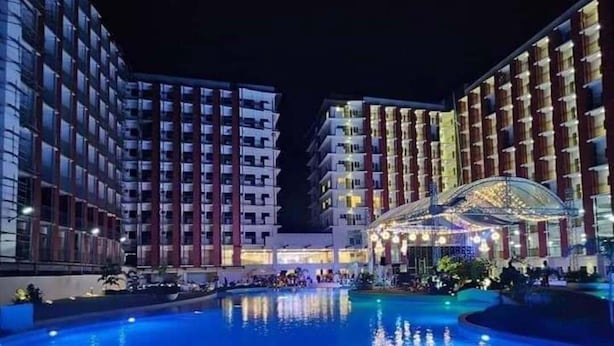 Une vue nocturne d’un hôtel doté d’une grande piscine, de plusieurs immeubles de grande hauteur et d’un espace couvert illuminé.