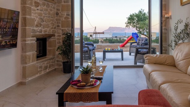 A living room with a stone fireplace, a sofa, a coffee table with a placemat, and a view of a slide and outdoor seating area. - The Hospitality of the 11 Brothers (kissamos)