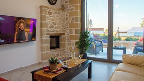 A living room with a stone fireplace, a television, a sofa, and a view of the outdoors. - The Hospitality of the 11 Brothers (kissamos)