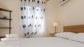 Une chambre d’hôtel avec un lit bien fait, une table de chevet avec une lampe, une étagère avec des objets décoratifs et une fenêtre avec des rideaux légers.