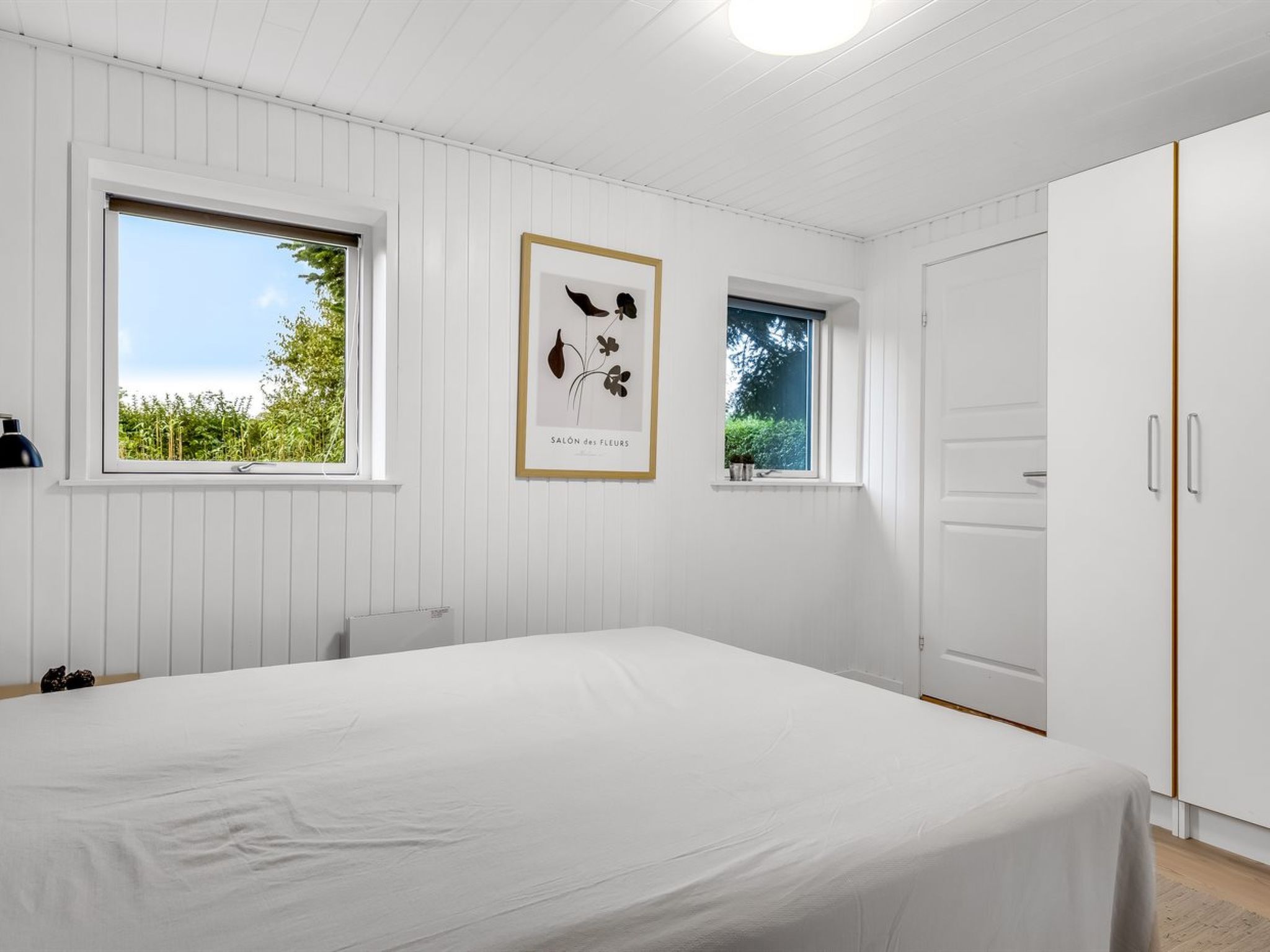 Une chambre à coucher avec un lit, deux fenêtres, une armoire et une estampe botanique encadrée accrochée au mur.