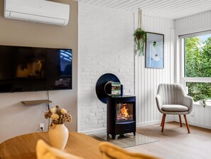 A modern living room with a flat-screen TV, a wood-burning stove, a comfortable armchair, and a large window with a view of greenery.