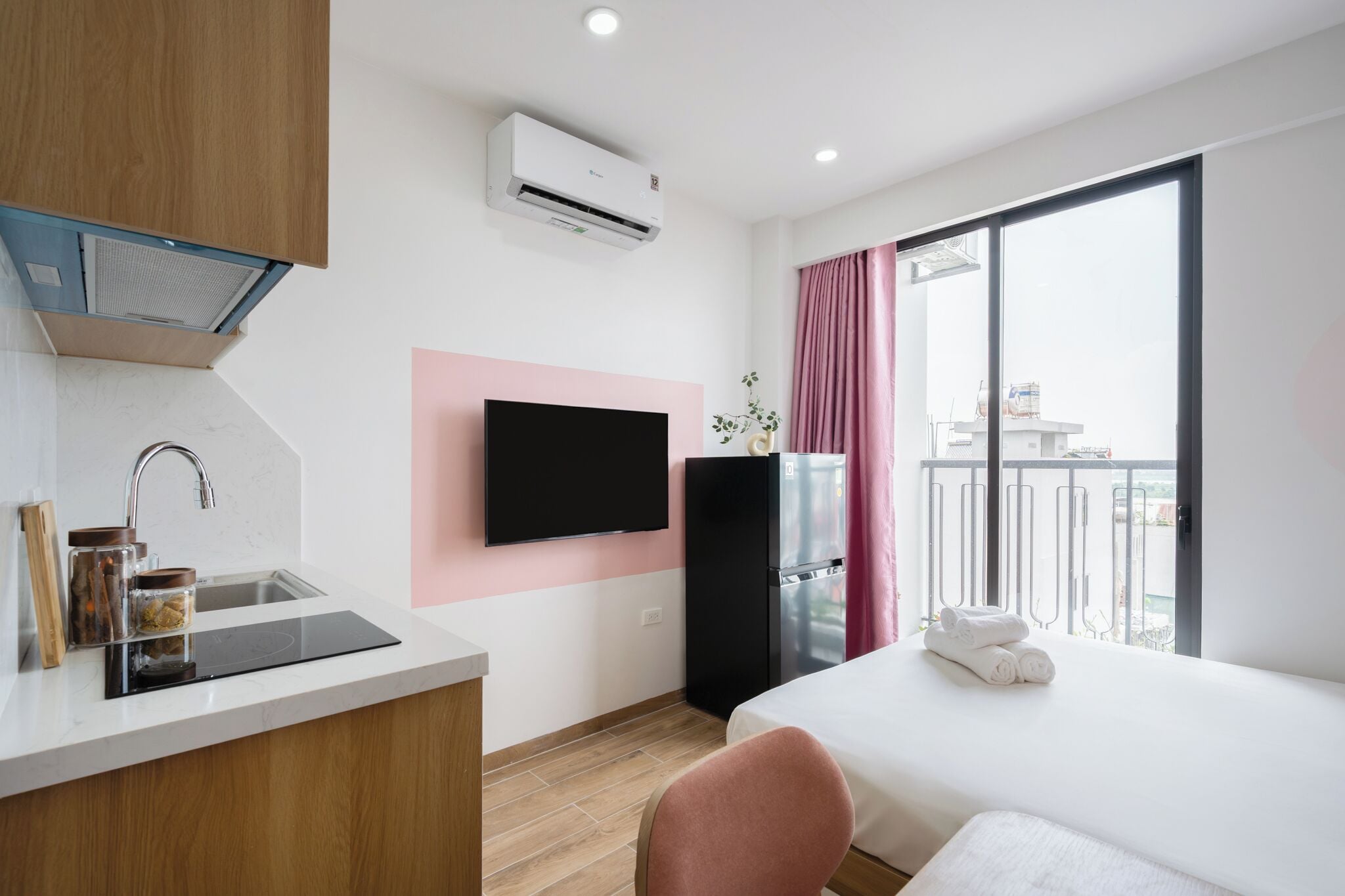 A modern hotel room with a kitchenette, a flat-screen TV, a minibar, a large window with a balcony, and a bed with rolled towels.