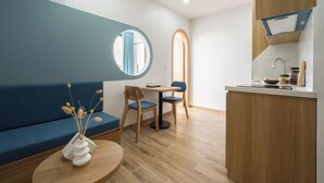 Quarto moderno com um sofá azul, uma mesa redonda com um vaso, uma pequena cozinha com pia e bancada, e um espelho na parede.