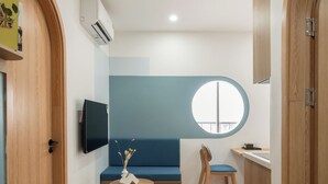 Quarto moderno com piso de madeira, sofá azul, janela redonda, televisão e uma pequena mesa com um vaso.
