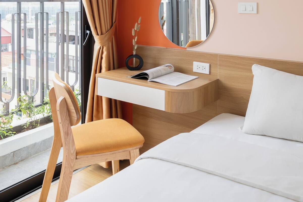 Habitación de hotel con cama, escritorio con espejo, una silla y vistas a la ciudad a través de amplias ventanas.