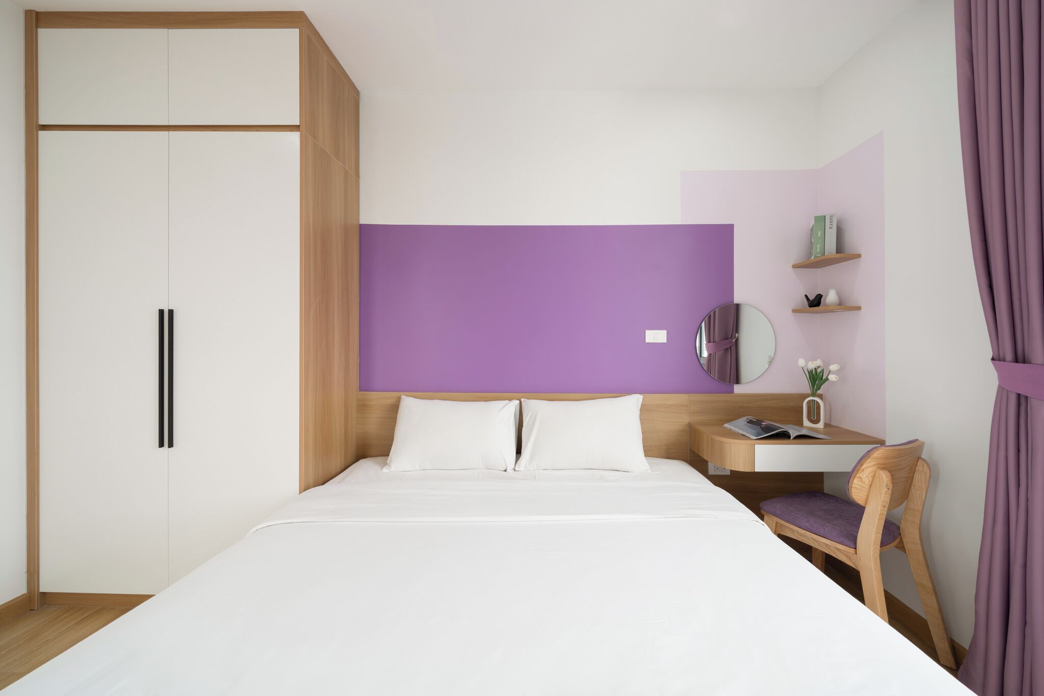 A bedroom with a bed, a wooden wardrobe, a desk with a chair, and a purple accent wall.