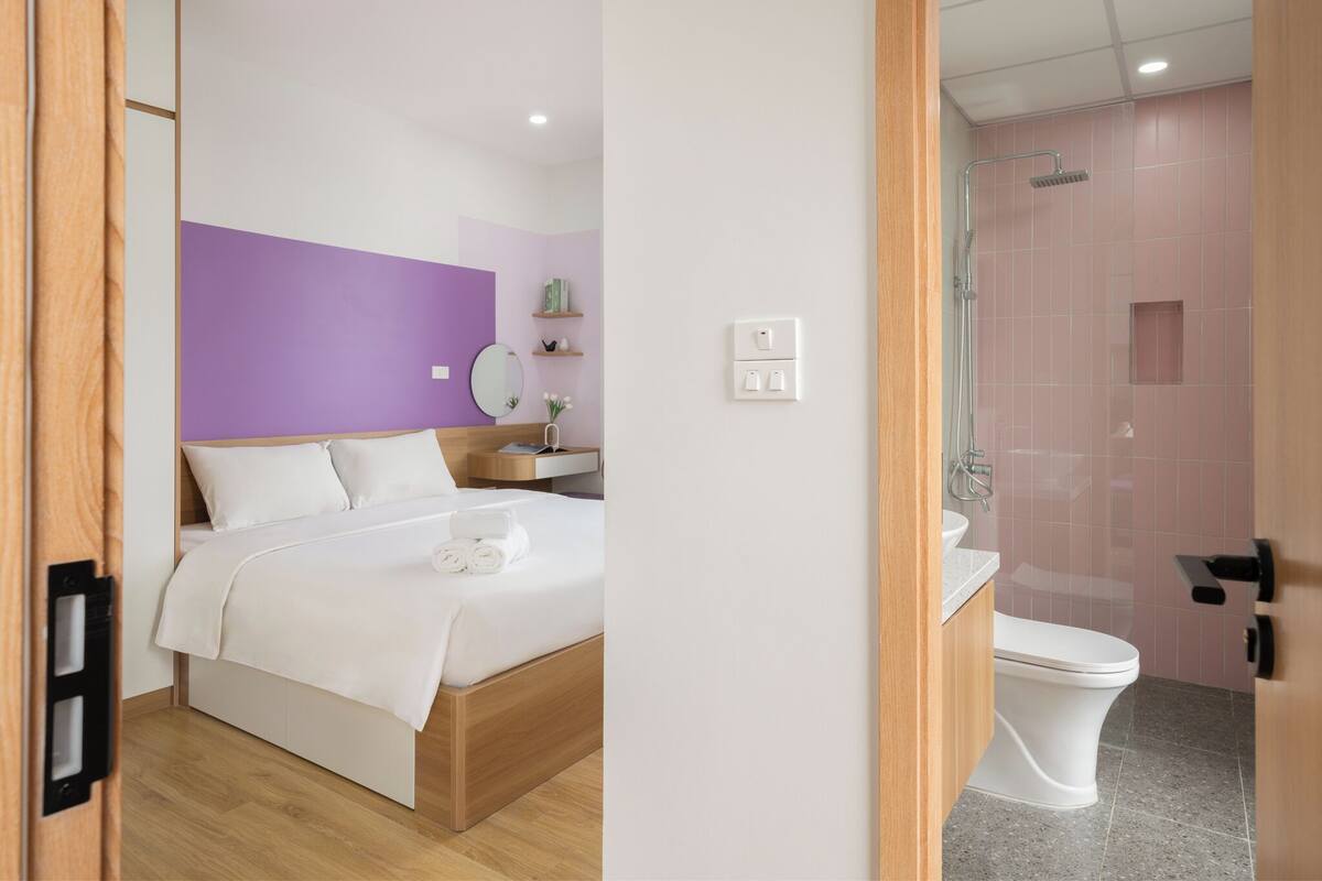 Una habitación de hotel moderna con una cama de madera, un baño con ducha y una pared de acento morada.
