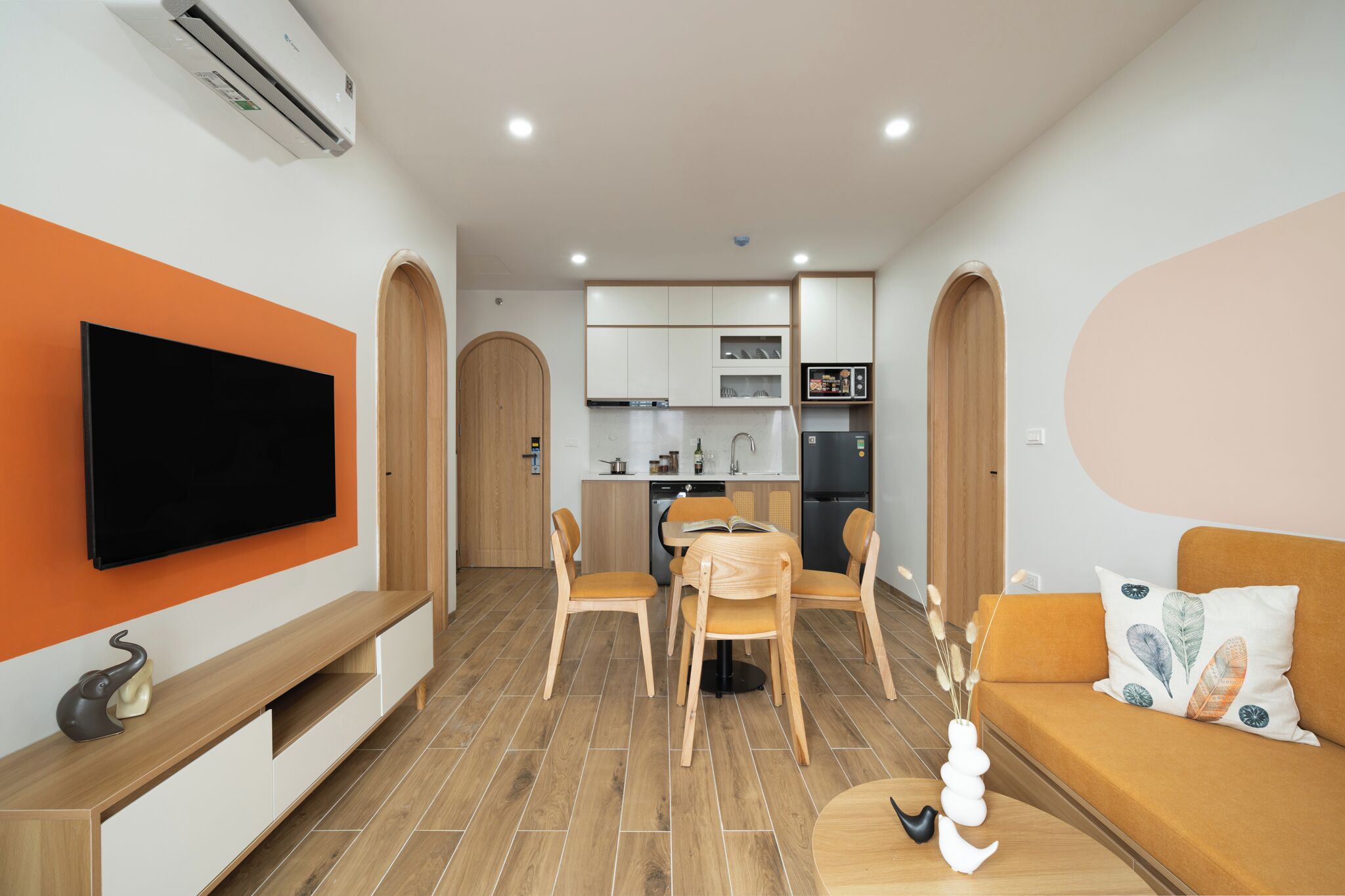 A modern living room with a flat-screen TV, wooden furniture, a sofa with decorative pillows, and a kitchen area in the background.