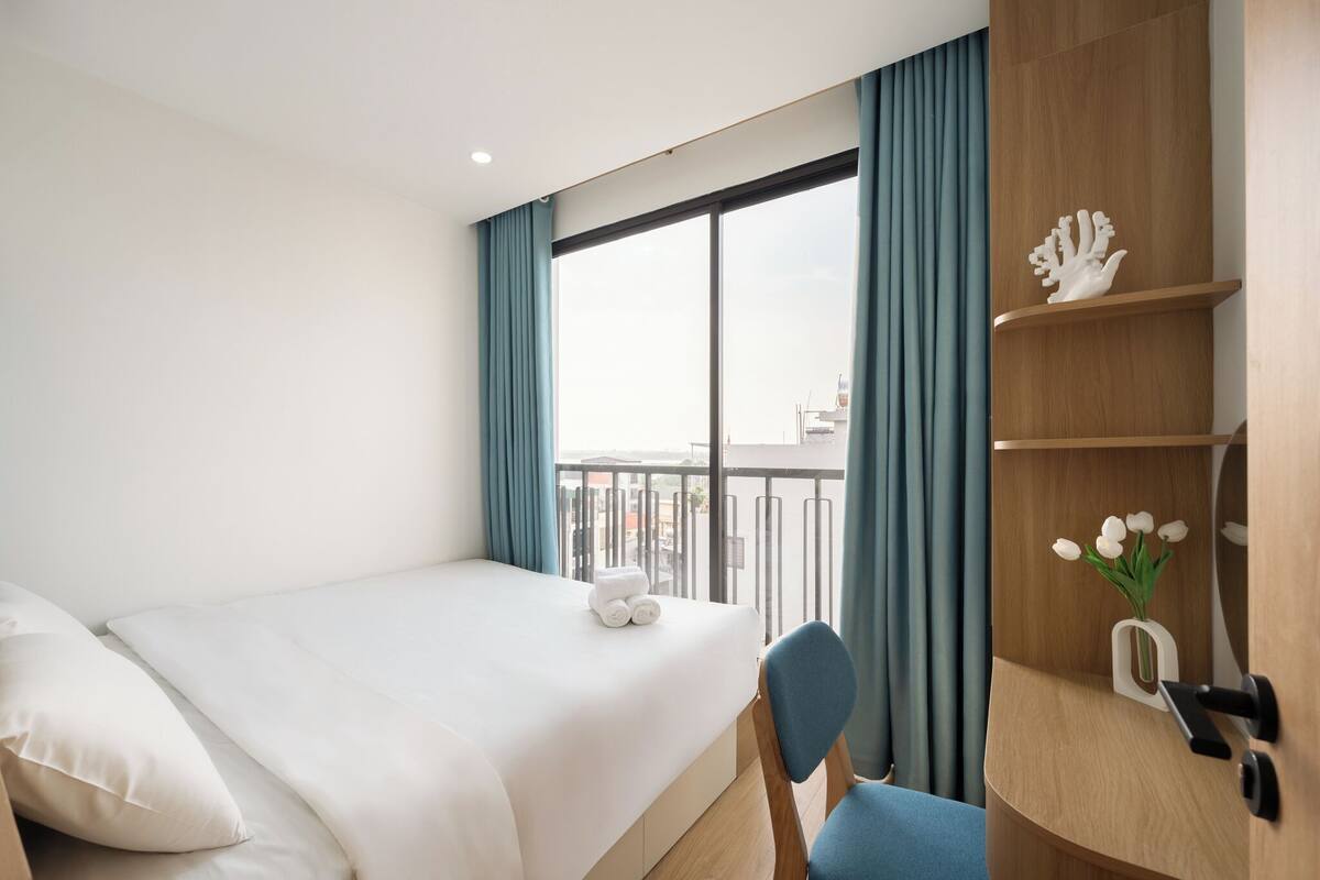 Habitación de hotel con cama, una silla, un escritorio con teléfono, un jarrón con flores y vistas a la ciudad a través de amplias ventanas.
