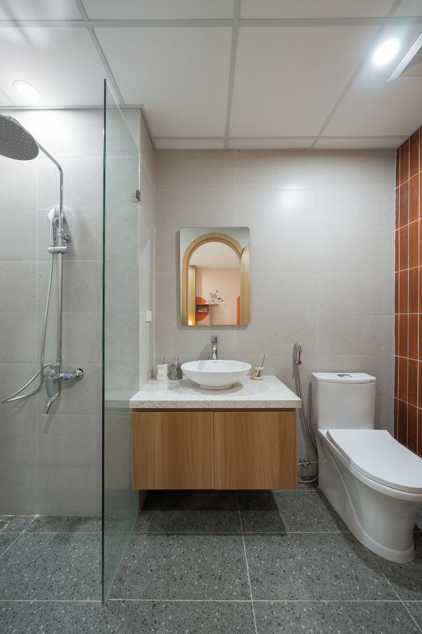 Un baño moderno con cabina de ducha de vidrio, un lavabo blanco con tocador de madera, un espejo redondo y un inodoro blanco.