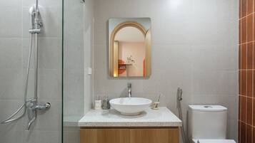 Un baño moderno con cabina de ducha de vidrio, un lavabo blanco con tocador de madera, un espejo redondo y un inodoro blanco.