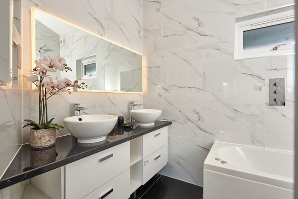 A modern bathroom with a marble backsplash, two sinks, a large mirror, and a bathtub. - Lakehurst Park (Milton Keynes)