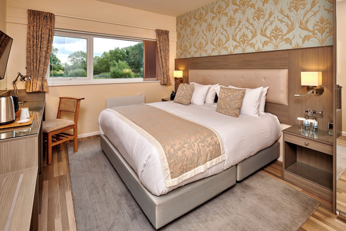 Quarto de hotel com uma cama grande, uma mesa com luminária, uma cadeira e vista para árvores pela janela.
