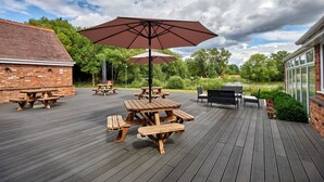 A wooden deck with picnic tables and umbrellas, surrounded by brick buildings and greenery.
