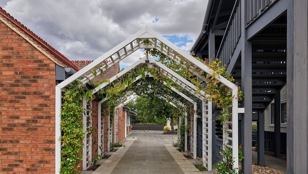 A covered walkway with brick walls and a tiled floor, flanked by greenery and a white trellis structure.