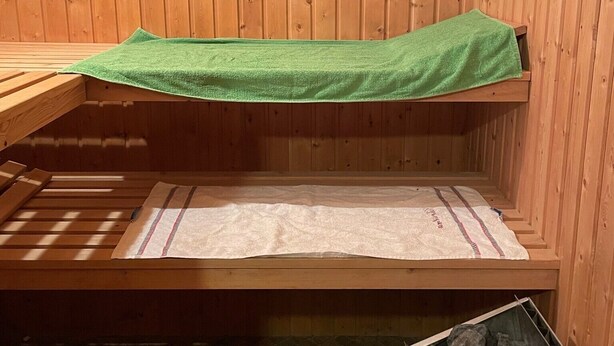 A sauna with wooden walls, a bench, a green towel, a bench with a white towel, and a container of rocks. - Entire Apartment in a Quiet Location (Oerlenbach)