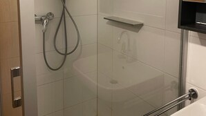 A shower with a handheld showerhead and a wall-mounted faucet, a glass shower enclosure, and a white sink with a mirror above it. - Entire Apartment in a Quiet Location (Oerlenbach)