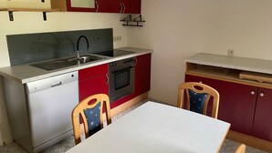 A compact kitchen with a dishwasher, oven, and sink, a dining table, and red cabinets. - Entire Apartment in a Quiet Location (Oerlenbach)