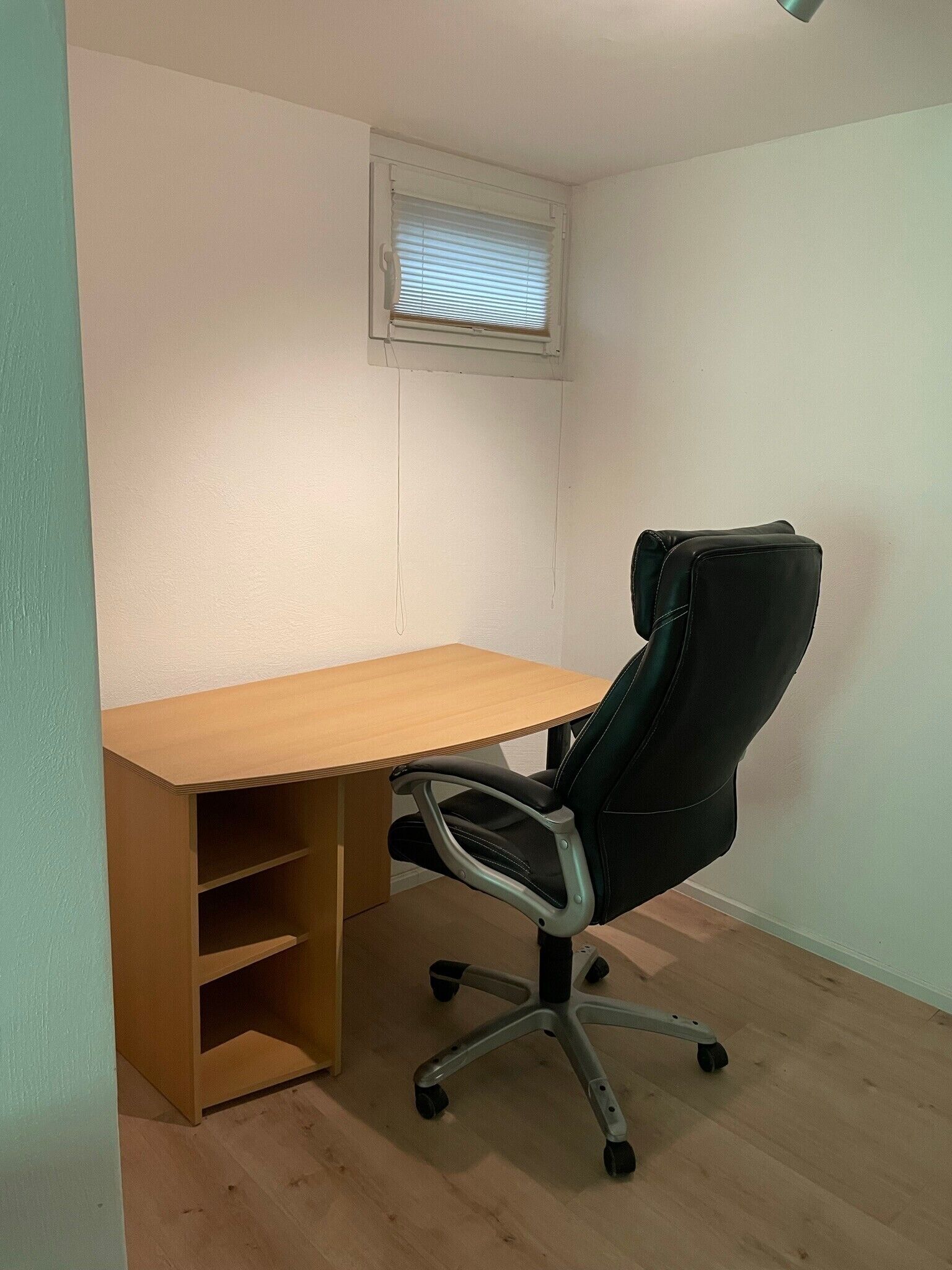Un pequeño espacio de oficina con un escritorio de madera, una silla de oficina negra y una ventana con persianas.