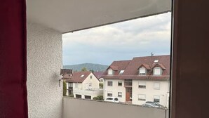 A balcony with a white chair and a small table, overlooking a residential area with houses and a hilly landscape.