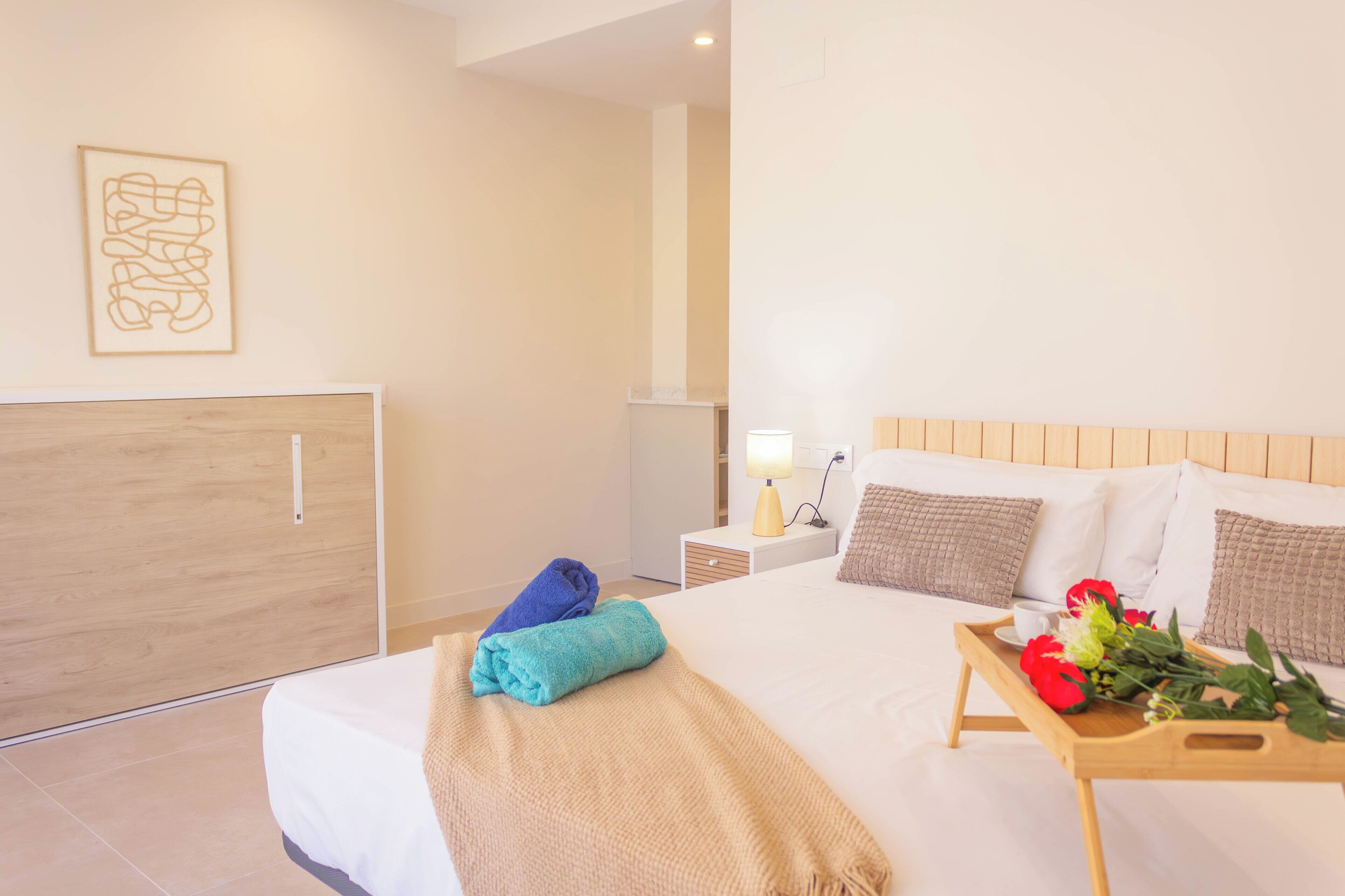 A hotel room with a bed, a bedside table with a lamp, a tray with flowers, and a wall art piece.