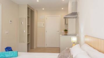 Una habitació d'hotel amb un llit, una taula de nit amb una làmpada, una petita cuina, un armari i una porta que dona a una altra habitació.