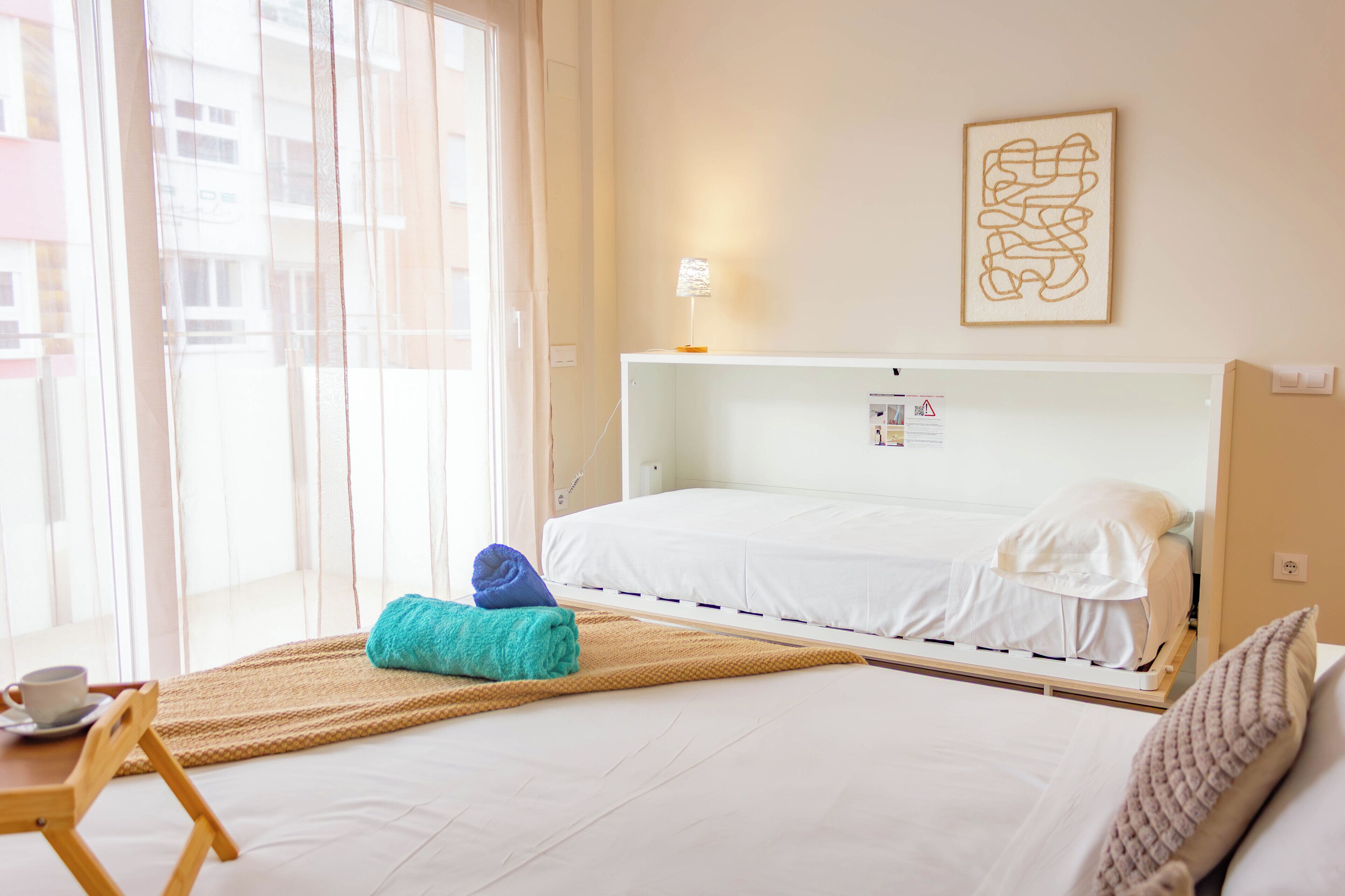 A hotel room with a bed, a small table with a cup, a window with curtains, and a wall-mounted lamp.