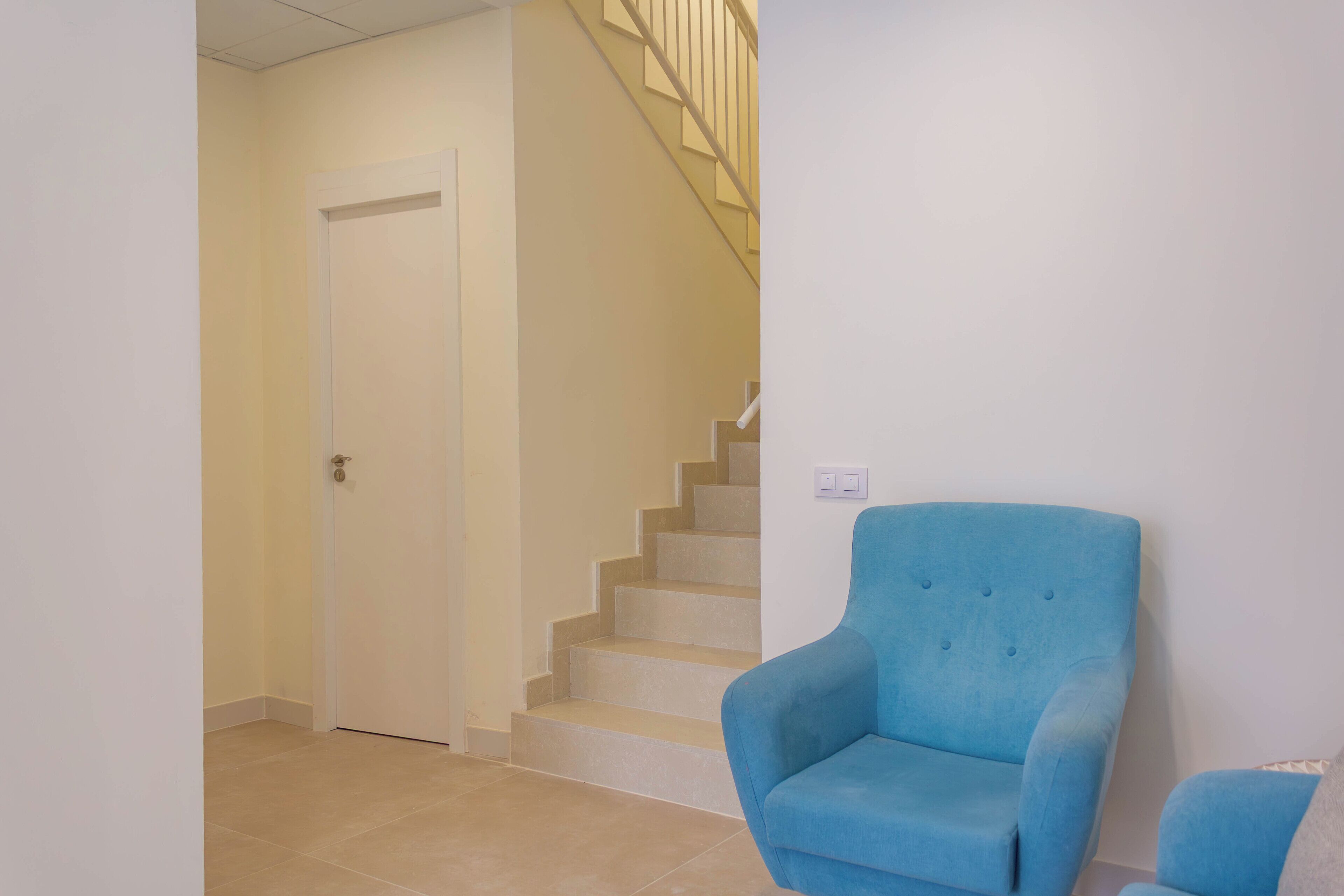 Een hal met een gesloten deur, een blauwe fauteuil en een trap met beige treden.