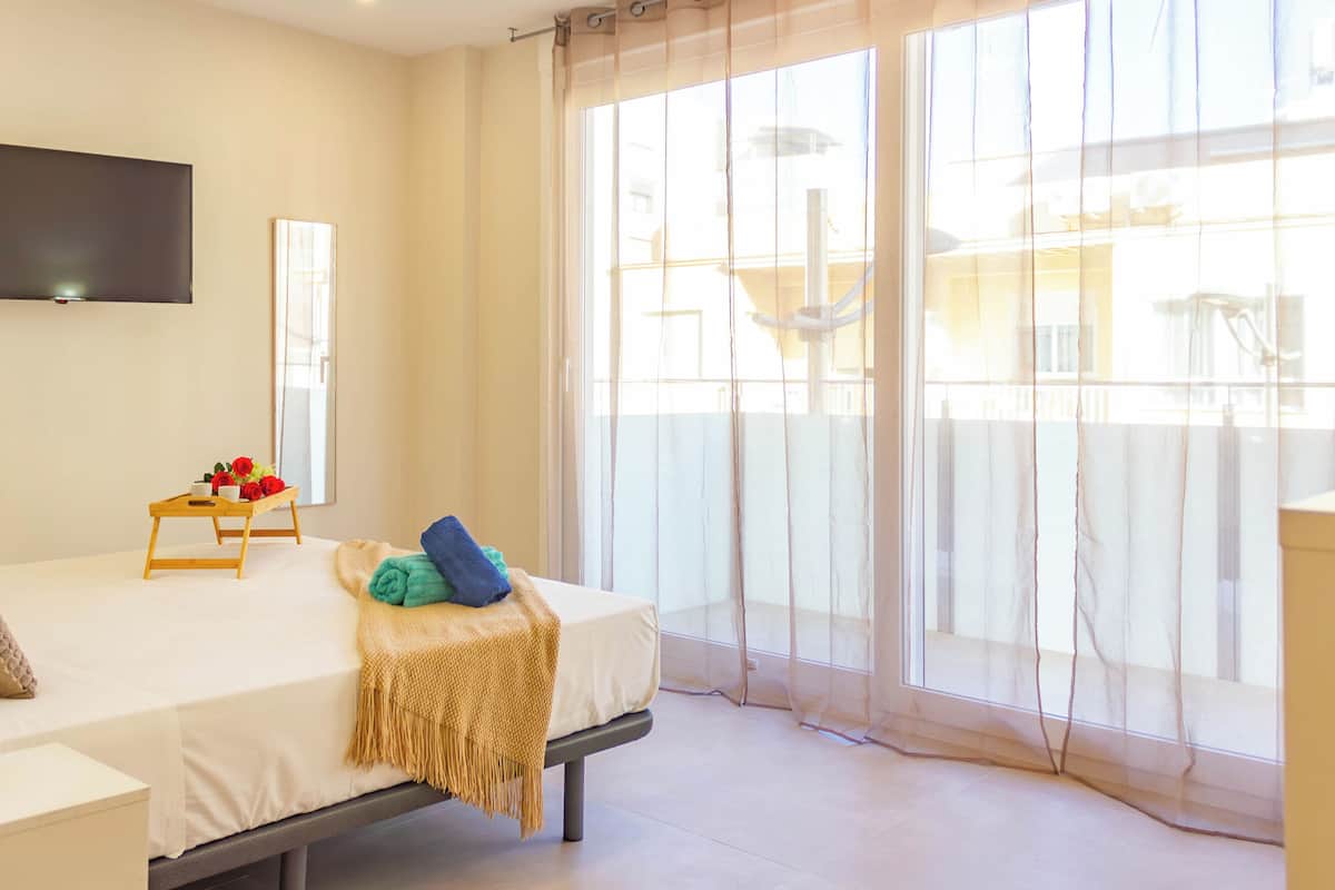 Un dormitorio con cama, mesita de noche, televisor montado en la pared y ventana con cortinas translúcidas.